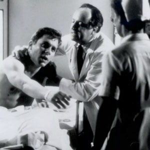 May include: A black and white photo of a man lying in a hospital bed, being examined by a doctor and a nurse.