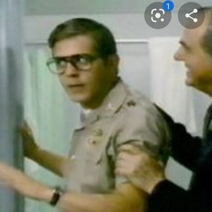 May include: A man in a khaki uniform with glasses looks nervously at another man in a dark suit. The man in the suit has his hand on the other man's shoulder.