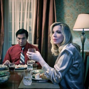 May include: A woman and a man sit at a table set for dinner. The woman is wearing a light gray satin blouse and has long blonde hair. The man is wearing a red shirt with a striped tie. There is a glass of wine in front of the woman and a glass of red wine in front of the man.