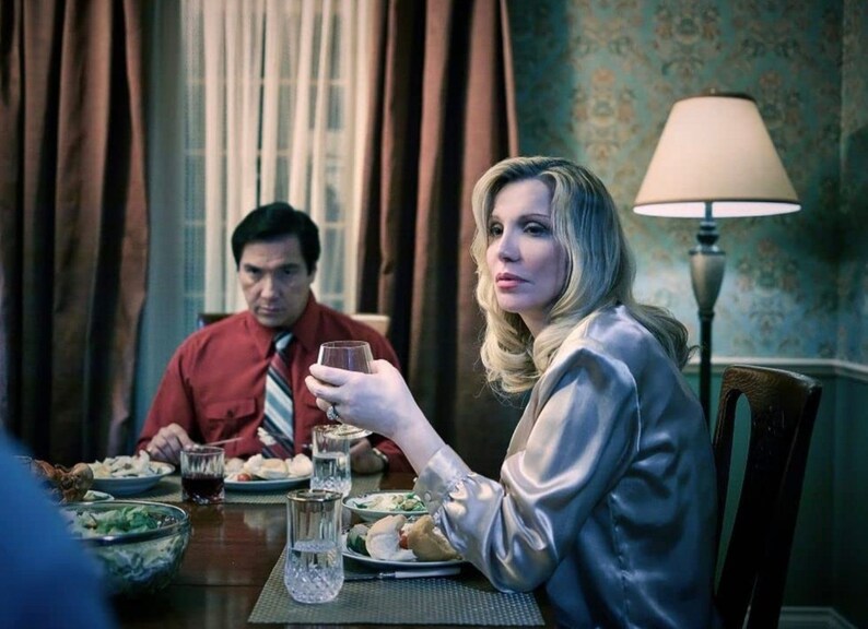 May include: A woman and a man sit at a table set for dinner. The woman is wearing a light gray satin blouse and has long blonde hair. The man is wearing a red shirt with a striped tie. There is a glass of wine in front of the woman and a glass of red wine in front of the man.