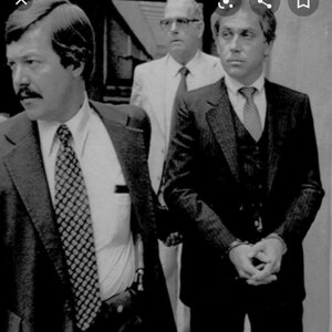 May include: Three men in suits, two wearing ties, one with a vest, standing in a courtroom. The man on the left has a mustache and is looking to the left. The man in the middle is wearing glasses and looking straight ahead. The man on the right is looking to the left.