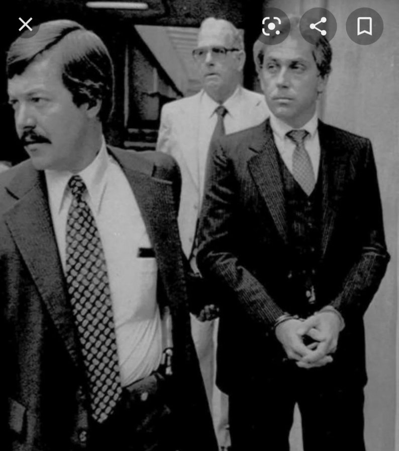 May include: Three men in suits, two wearing ties, one with a vest, standing in a courtroom. The man on the left has a mustache and is looking to the left. The man in the middle is wearing glasses and looking straight ahead. The man on the right is looking to the left.