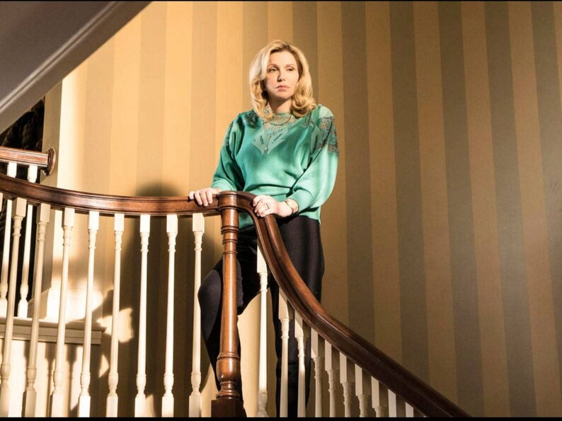 May include: A woman in a green sweater and black pants stands on a staircase with a wooden railing. The background is a striped wall.