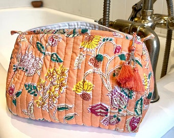 Op de afbeelding: Een roze quilted toilettas met een bloemenprint en een kwast. De tas ligt op een wit aanrecht in een badkamer.