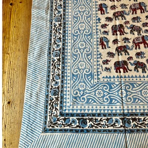 May include: A white cotton tablecloth with a blue and black border. The center of the tablecloth features a repeating pattern of elephants in red, black, and white.