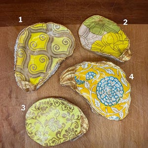 May include: Four decorative oyster shells with gold-painted edges. The shells feature various fabric patterns in yellow, green, and blue. Designs include floral and geometric patterns. The shells are arranged on a wooden surface.