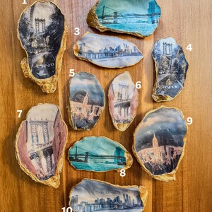 May include: Ten oyster shell trinket dishes, each with a different image of New York City landmarks. The shells have gold edges and are displayed on a wooden surface. The images include bridges and city skylines.