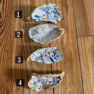 May include: Four decorative oyster shell dishes with gold-painted edges. The shells are adorned with floral patterns in various colors, including pink, blue, and white. The shells are arranged on a wooden surface.