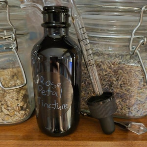May include: A dark glass bottle with a black label that reads "Rose Petal Tincture". The bottle is sitting on a wooden surface next to a glass jar filled with dried lavender.