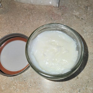 May include: A clear glass jar filled with a white, creamy substance. The jar's lid is off to the side, revealing a red and silver rim. The jar sits on a light brown surface, with a bag of white crystals in the background.