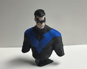 May include: A sculpted bust of a character with dark hair, wearing a black mask over the eyes and a black suit featuring a prominent blue chevron design across the chest and shoulders. The figure is depicted from the chest up with a neutral expression.