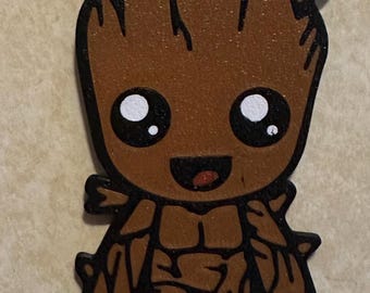 May include: A brown wooden keychain shaped like a small tree creature with a textured bark appearance, large black eyes with white highlights, and a smiling mouth. The figure features dark outlines and a metal loop at the top for attachment.