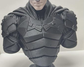 May include: A matte black bust of the character Batman, featuring a detailed cowl with pointed ears, a textured chest plate with segmented armor plating, and a cape draped over the shoulders. The figure displays a neutral facial expression.