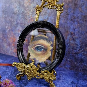 May include: A single blue eye painted on canvas is framed in a black circular frame with ornate gold trim. The frame is on a gold easel.