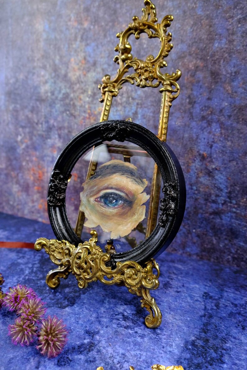 May include: A single blue eye painted on canvas is framed in a black circular frame with ornate gold trim. The frame is on a gold easel.