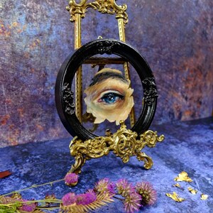 May include: A blue eye painted on a canvas, framed in a black oval frame and displayed on a gold easel. The easel is on a blue textured surface with gold leaf accents.
