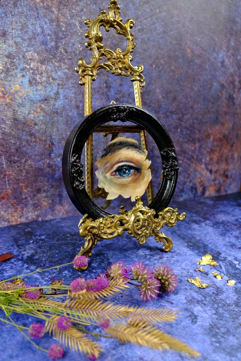 May include: A blue eye painted on a canvas, framed in a black oval frame and displayed on a gold easel. The easel is on a blue textured surface with gold leaf accents.