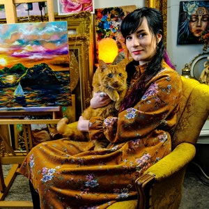 May include: A woman in a floral dress holds a ginger cat in her lap. She is sitting in a yellow armchair in a room with paintings on the walls.