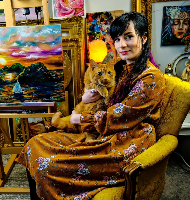 May include: A woman in a floral dress holds a ginger cat in her lap. She is sitting in a yellow armchair in a room with paintings on the walls.