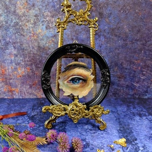 May include: A blue eye painted on a canvas, framed in a black oval frame and displayed on a gold easel. The background is a blue and purple textured surface.