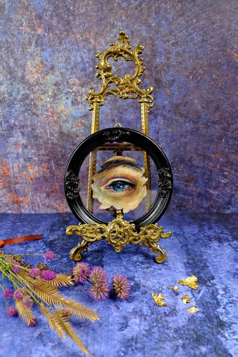 May include: A blue eye painted on a canvas, framed in a black oval frame and displayed on a gold easel. The background is a blue and purple textured surface.
