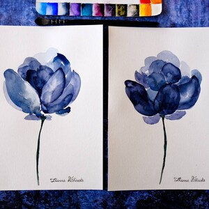 May include: Two watercolor paintings of blue flowers on white paper. Each flower has a dark blue center and lighter blue petals. A green stem supports each flower. The artist's signature is at the bottom. Watercolor paints and a brush are visible.