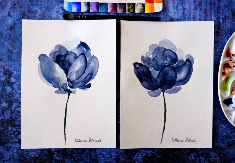 May include: Two watercolor paintings of blue flowers on white paper. Each flower has a dark blue center and lighter blue petals. A green stem supports each flower. The artist's signature is at the bottom. Watercolor paints and a brush are visible.