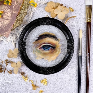 May include: A close-up of a framed painting of a blue eye. The eye is painted in a realistic style and is surrounded by a light brown skin tone. The frame is black and has a decorative design.