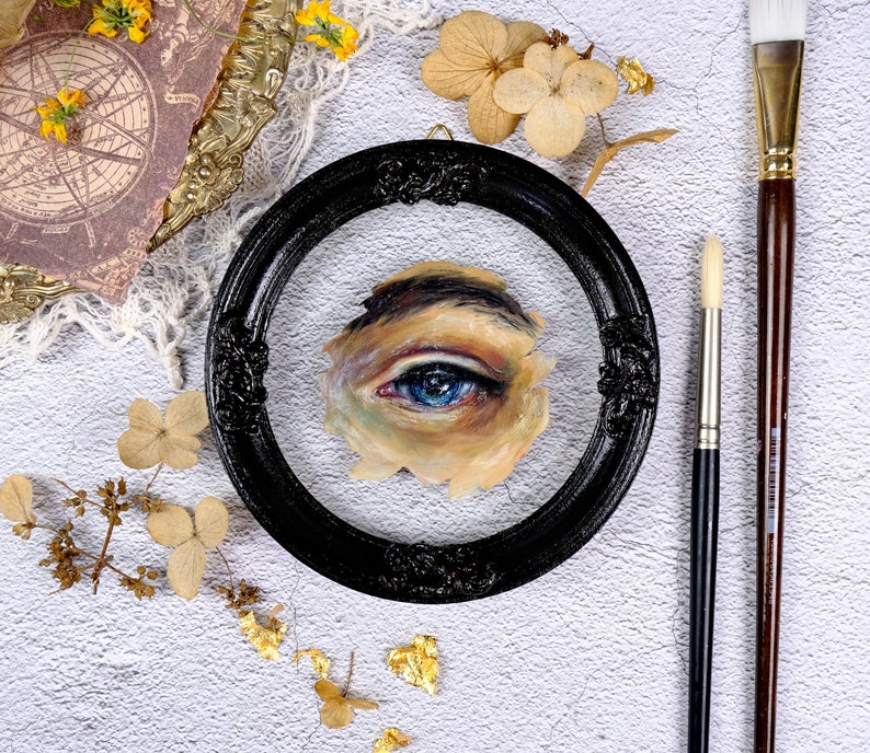 May include: A close-up of a framed painting of a blue eye. The eye is painted in a realistic style and is surrounded by a light brown skin tone. The frame is black and has a decorative design.