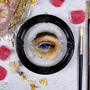 May include: A close-up of a framed painting of a blue eye. The eye is painted in a realistic style and is surrounded by skin tones. The frame is black and ornate.