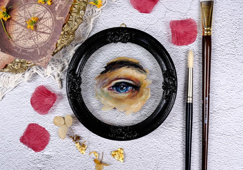 May include: A close-up of a framed painting of a blue eye. The eye is painted in a realistic style and is surrounded by skin tones. The frame is black and ornate.