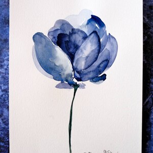 May include: Watercolor painting of a blue flower with a dark green stem. The flower has multiple layers of blue petals, with lighter shades in the center. The artist's signature, "Dhanna Vitkovska," is in the lower right corner.