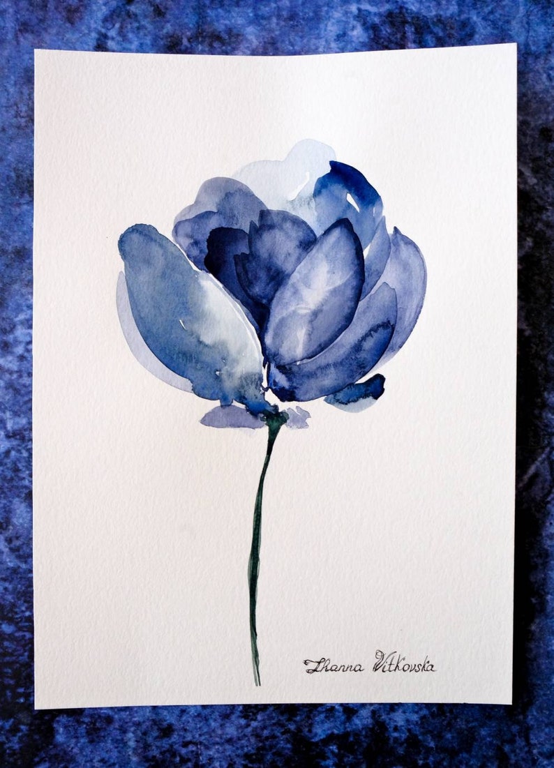 May include: Watercolor painting of a blue flower with a dark green stem. The flower has multiple layers of blue petals, with lighter shades in the center. The artist's signature, "Dhanna Vitkovska," is in the lower right corner.