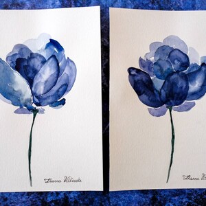 May include: Two watercolor paintings of blue flowers on white paper. Each flower has multiple layers of blue petals and a long green stem. The artist's signature, "Zianna Vitkouska", is written in cursive at the bottom of each painting.
