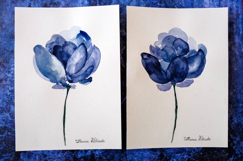 May include: Two watercolor paintings of blue flowers on white paper. Each flower has multiple layers of blue petals and a long green stem. The artist's signature, "Zianna Vitkouska", is written in cursive at the bottom of each painting.