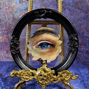 May include: A close-up of a painted eye with blue iris and brown eyelashes, framed in a black oval frame and displayed on a gold easel.
