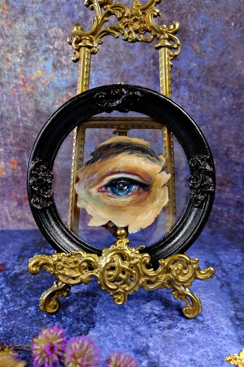 May include: A close-up of a painted eye with blue iris and brown eyelashes, framed in a black oval frame and displayed on a gold easel.