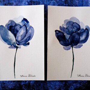 May include: Two watercolor paintings of blue flowers on white paper. Each painting features a single flower with a long stem. The flowers are in shades of blue, with darker hues in the center. The artist's signature is at the bottom.