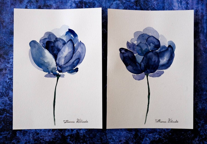 May include: Two watercolor paintings of blue flowers on white paper. Each painting features a single flower with a long stem. The flowers are in shades of blue, with darker hues in the center. The artist's signature is at the bottom.