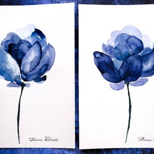 May include: Two watercolor paintings of blue flowers on white paper. Each flower has multiple layers of blue petals and a thin green stem. The artist's signature, "Zanna Vitkovska," is at the bottom of each painting.