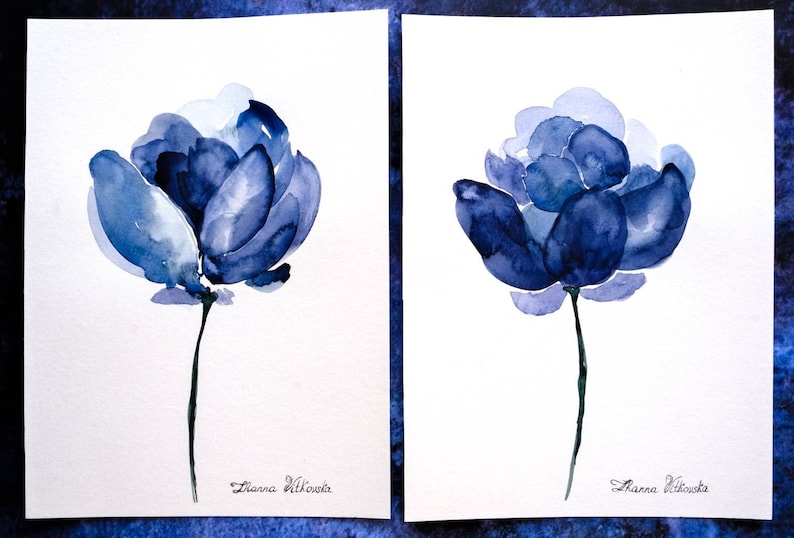 May include: Two watercolor paintings of blue flowers on white paper. Each flower has multiple layers of blue petals and a thin green stem. The artist's signature, "Zanna Vitkovska," is at the bottom of each painting.