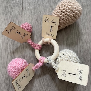 May include: A wooden baby teether with pink, beige, and white crocheted balls attached. The teether has tags with the text "Day 1 First Month", "Day 5 Walnut", "Week 1 April".