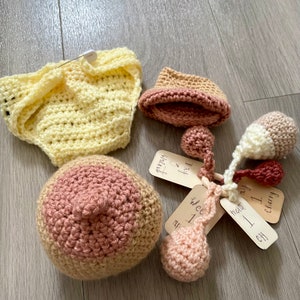 May include: A set of crocheted baby items in pink, yellow, and cream colors. The set includes a diaper cover, a hat, a rattle, and a set of milestone cards with the text "Day 2 & Infant", "Week 1", "Month 1", and "Cherry 7 Day".