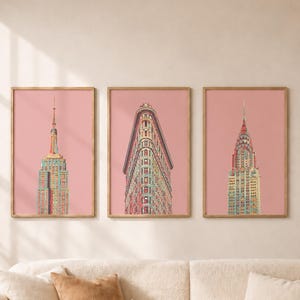 May include: A set of three framed vertical wall art prints featuring illustrations of the Empire State Building, the Flatiron Building, and the Chrysler Building, each set against a solid dusty pink background in light wood frames.