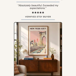 May include: A framed vertical wall art print titled NEW YORK CITY featuring a stylized illustration of the Brooklyn Bridge and city skyline in pink and blue tones, displayed above a dark wood sideboard with books, a lamp, and a small vase.
