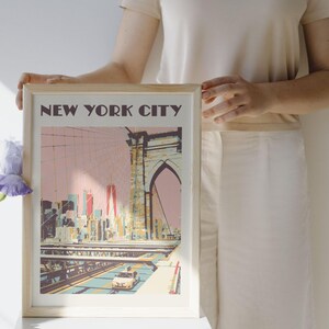 A framed art print featuring a stylized illustration of the Brooklyn Bridge and New York City skyline in shades of pink, yellow, and blue. The text NEW YORK CITY is printed in dark brown at the top of the print, which shows a yellow taxi.