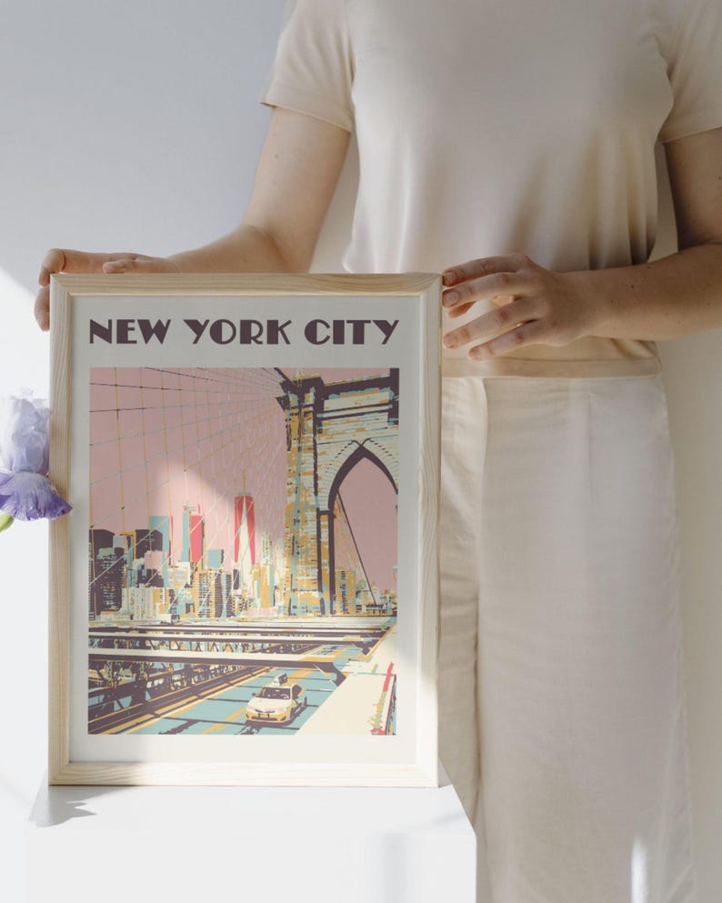 A framed art print featuring a stylized illustration of the Brooklyn Bridge and New York City skyline in shades of pink, yellow, and blue. The text NEW YORK CITY is printed in dark brown at the top of the print, which shows a yellow taxi.