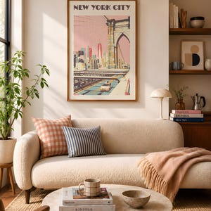 Retro travel poster print of the Brooklyn Bridge, New York City. Illustrated in warm pinks, yellow, and teals with a vintage flat-graphic style. The Manhattan skyline rises in the background beneath a blush sky. Shown framed in a walnut frame above a cream boucle sofa styled with gingham and striped cushions in a warm, sunlit living room. Coffee table styled with Kinfolk books and a stoneware mug.