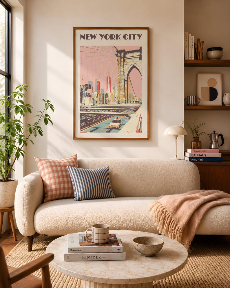 Retro travel poster print of the Brooklyn Bridge, New York City. Illustrated in warm pinks, yellow, and teals with a vintage flat-graphic style. The Manhattan skyline rises in the background beneath a blush sky. Shown framed in a walnut frame above a cream boucle sofa styled with gingham and striped cushions in a warm, sunlit living room. Coffee table styled with Kinfolk books and a stoneware mug.