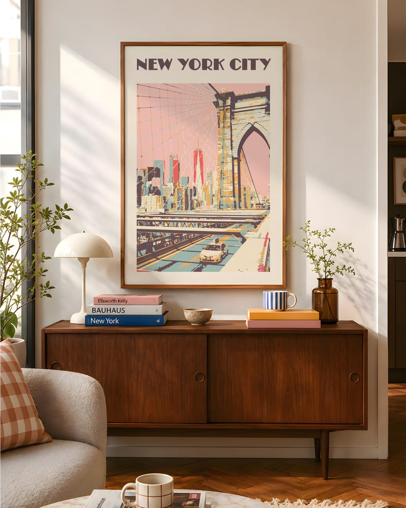 Retro travel poster print of the Brooklyn Bridge, New York City. Illustrated in warm pinks, yellow, and teals with a vintage flat-graphic style. The Manhattan skyline rises in the background beneath a blush sky. Shown framed in a walnut frame above a mid-century modern credenza in a warm, sunlit living room. Styled with art books, a mushroom lamp, and green branches.